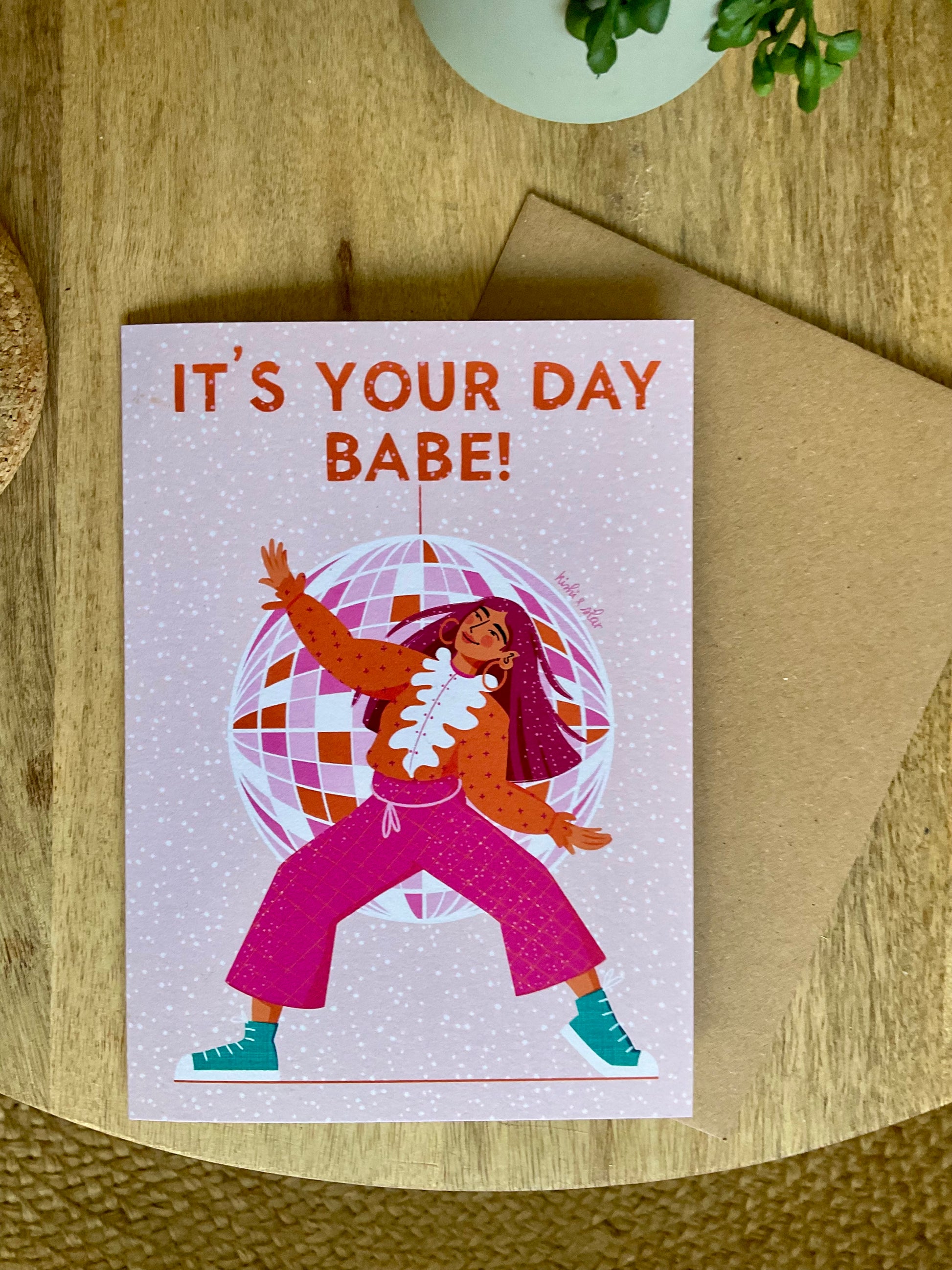 Greeting card with illustration of a person and text 'It's Your Day Babe!' on a wooden surface.
