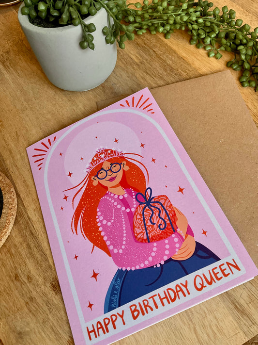 Greeting card with illustration of a woman and 'Happy Birthday Queen' text on a wooden surface with a plant.